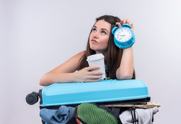 Thoughtful young traveler woman holding alarm clock and travelling plastic coffee cup with suitcase on isolated white wall