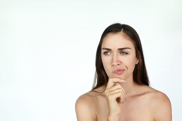 Thoughtful young woman holds fingers on her chin standing on white background