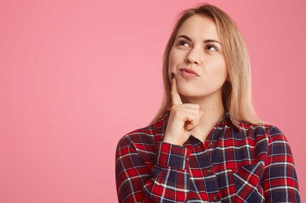 thoughtful young woman holds fore finger near chin, focused up, daydreams about something