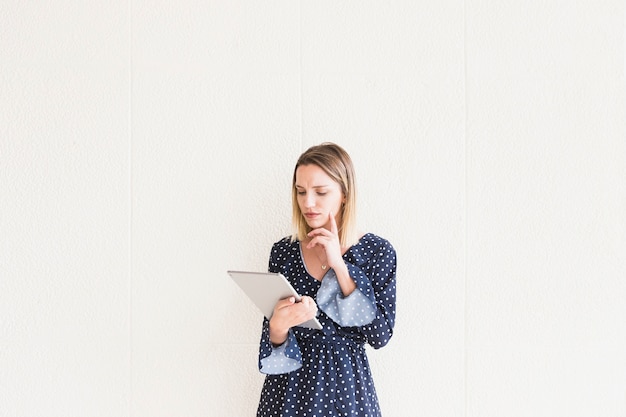 Thoughtful young woman looking at digital tablet in front of wall