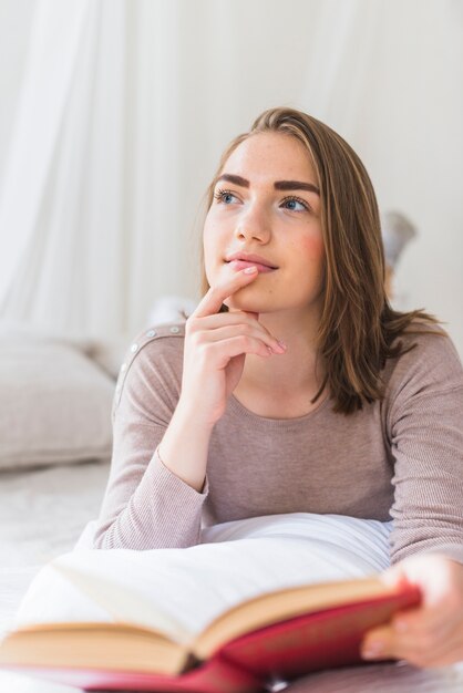 Thoughtful young woman lying on bed holding book