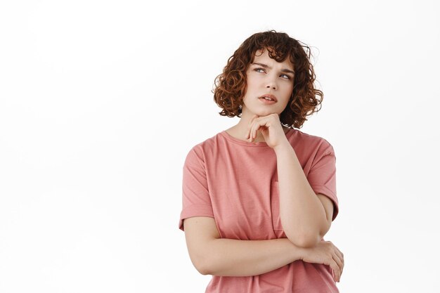 Thoughtful young woman making choice. Student thinking and looking up serious, thinking, deciding something, standing in t-shirt against white background