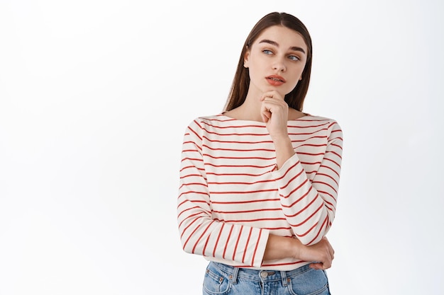 Thoughtful young woman making decision, make up her mind, looking aside at logo product, deciding, thinking about copyspace, standing over white background