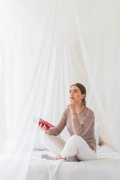 Free photo thoughtful young woman sitting on bed holding book in hand