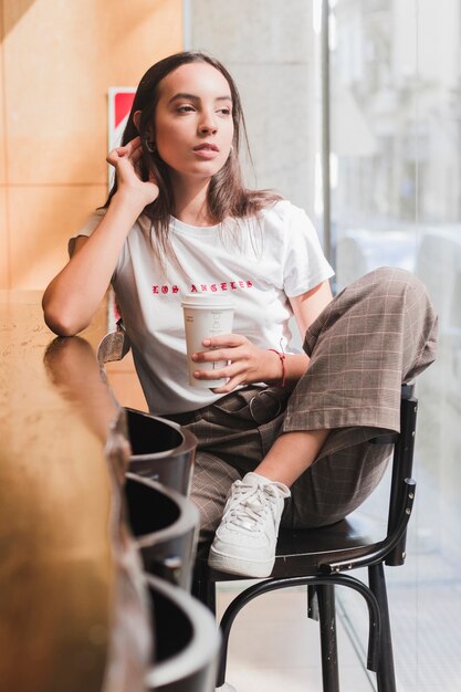 Thoughtful young woman sitting on chair in cafe holding the disposable coffee cup