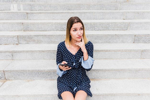 Thoughtful young woman sitting on staircase holding cellphone