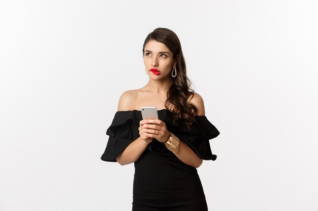 Thoughtful young woman using smartphone and thinking, looking up and pondering, standing over white background