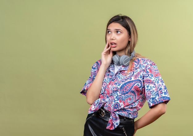 A thoughtful young woman wearing paisley printed shirt in headphones thinking with hand on face while looking up