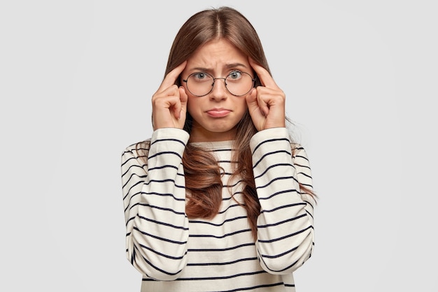 Thoughtful young woman with bad memory, keeps fingers on temples, has dissatisfied expression, dressed in striped casual sweater, isolated over white wall. People and thoughts concept.