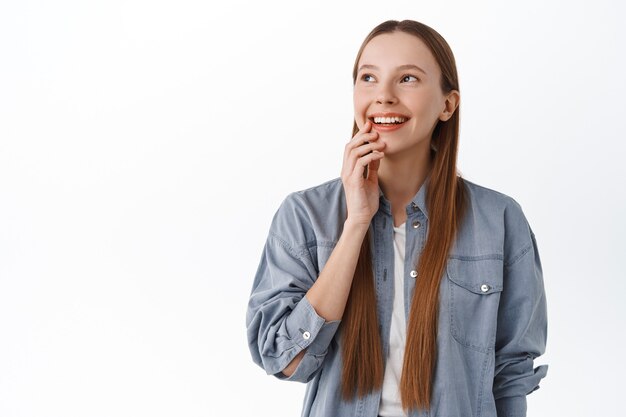 Thoughtful young woman with long hair thinking, smiling pleased and looking aside at copy space promotional text, imaging something, standing against white wall