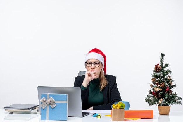Задумчивая молодая женщина в шляпе санта-клауса сидит за столом с рождественским деревом и подарком на нем и указывает вверх на белом фоне