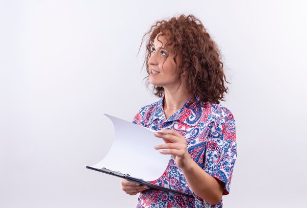 Thoughtful young woman with short curly hair  in colorful shirt holding clipboard with blank pages looking up 