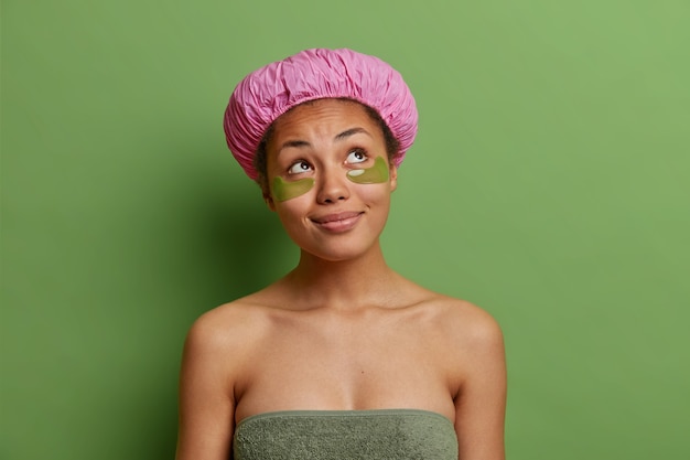 Thoughtful young woman wrapped in bath towel applies hydrogel patches under eyes wears bath hat isolated over vivd green wall