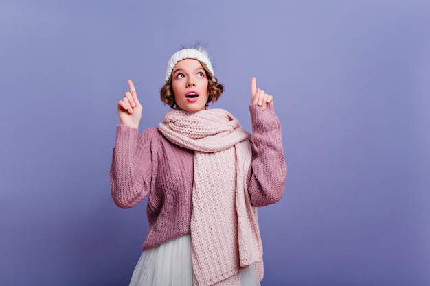 Thoughtfully short-haired girl in cute hat looking up with mouth open. Carefree female model posing in winter accessories on purple wall.