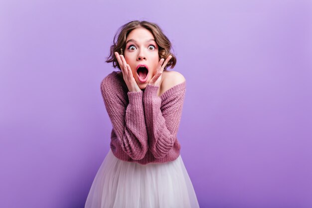 Thoughtfully short-haired girl in cute hat looking up with mouth open. Carefree female model posing in winter accessories on purple wall.