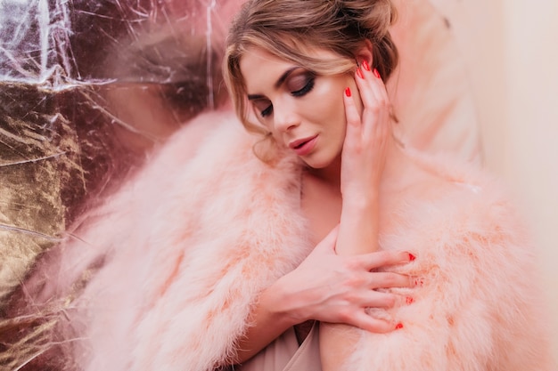 Thoughtfully stylish girl with dark make-up touching ear while posing on glitter silver wall. Amazing young woman with trendy hairstyle and manicure thinking about something enjoyable and looking down