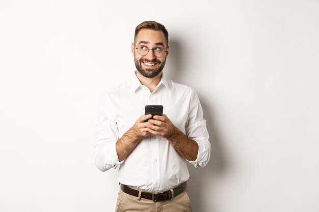 Thouthful handsome manager using mobile phone and thinking about answering message, looking at upper left corner and smiling, standing  