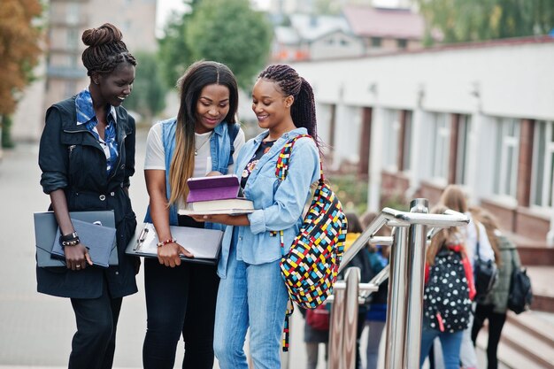 Three african students female posed with backpacks and school items on yard of university and look at tablet