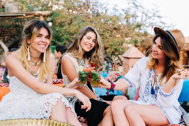 Three amazing girls in stylish vintage clothes having fun in outdoor cafe and drinking cocktails. Group of friends celebrating vacation and posing outside, holding glasses with beverage and laughing