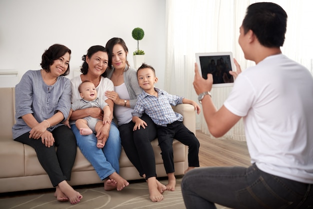 Три азиатские дамы с маленьким мальчиком и ребенком, сидя на диване и мужчина фотографировать на планшете