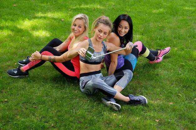 세 명의 매력적인 스포티 여성이 잔디밭에 앉아서 운동 후 셀카를 찍고 있습니다.