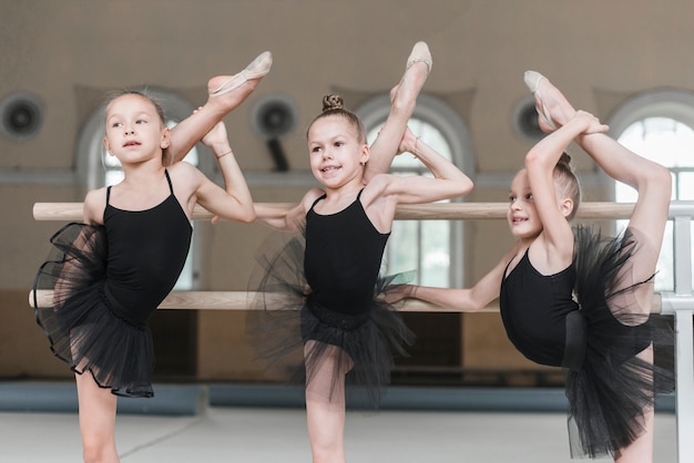 Three ballerina girls stretching their legs on barre in dance studio