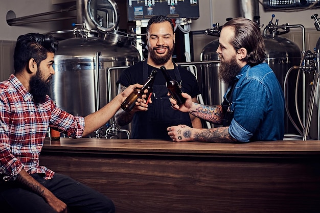 Three bearded interracial friends drink craft beer and talk in a brewery. Two hipster workers in aprons drink beer with their friend in a brewery factory. Old friends gathered to drink beer and chat.