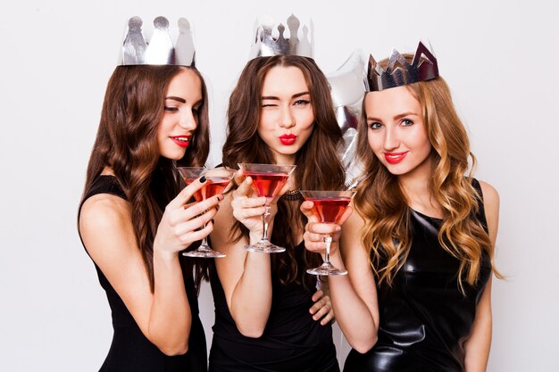 Three beautiful  elegant women celebrate hen-party and drinking  cocktails.  Best friends wearing black  evening dress ,crown  on head and clink glasses. Bright make up, red lips. Inside.