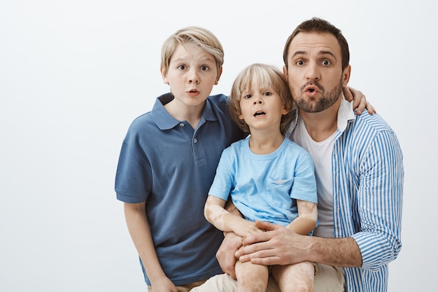 Three beautiful family members standing over grey wall, making surprised and amazed expressions while hugging