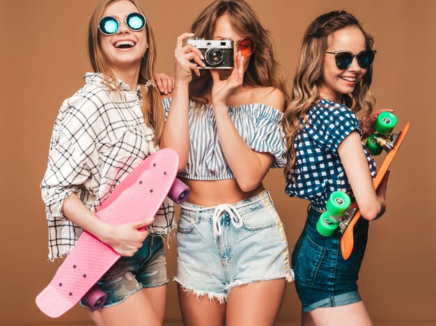 화려한 페니 스케이트 보드와 세 아름 다운 세련 된 웃는 여자. 여름 체크 무늬 셔츠 옷 포즈에 여자입니다. 레트로 사진 카메라로 사진 찍기