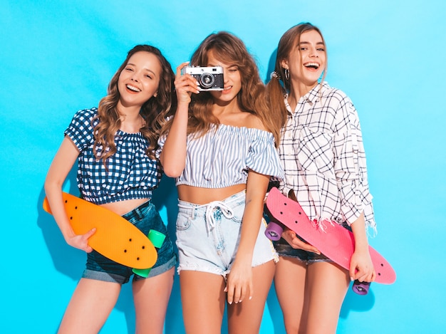 カラフルなペニースケートボードを持つ3つの美しいスタイリッシュな笑顔の女の子。夏の市松模様のシャツの女性。レトロな写真カメラで写真を撮る