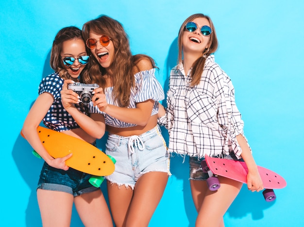 Three beautiful stylish smiling girls with colorful penny skateboards. Women in summer checkered shirt clothes. Taking pictures on retro photo camera