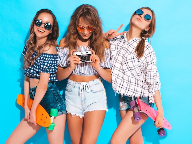 Tre belle ragazze sorridenti alla moda con i pattini del penny in occhiali da sole. le donne in abiti camicia estiva a scacchi. scattare foto con la retro macchina fotografica