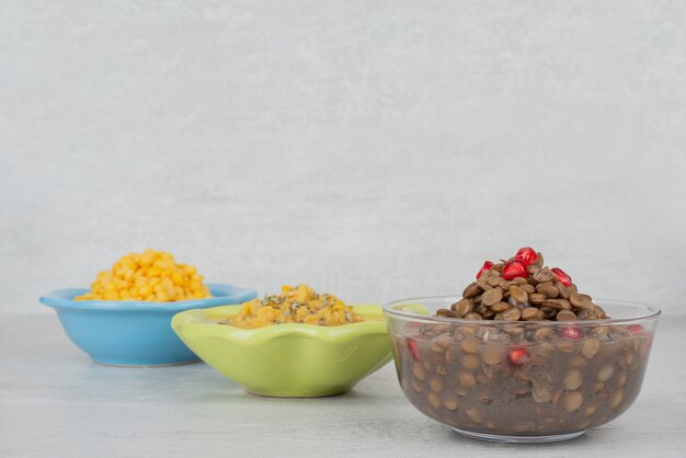 Three bowls of boiled corn, soup and beans on white surface.