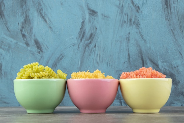 Three bowls of colorful fusilli on marble surface.