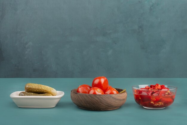 Three bowls of pickled red pepper, tomatoes and cucumbers on blue table.