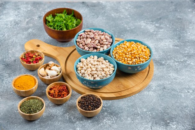 Three bowls of various beans and corns with spices on wooden board.