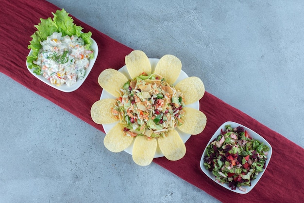 Three bowls of various salads garnished with lettuce leaf and potato chips on marble surface