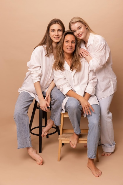 Three caucasian women of different ages in white shirts and jeans look at camera on beige background Concept of relatives
