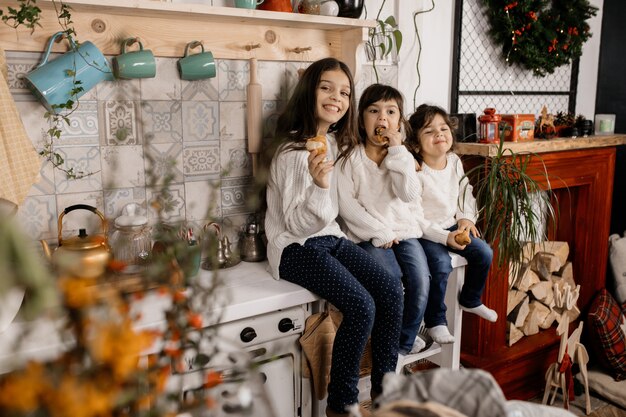 Three charming little girls in white sweaters and blue jeans play on an old-fashioned kitchen