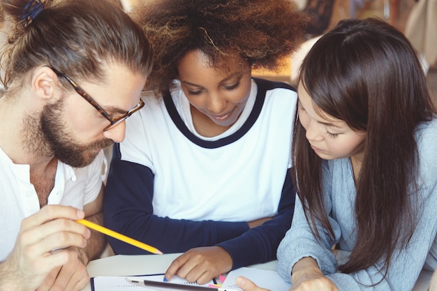 Three college students working on home assignment, sitting at cafeteria, making research, looking for required information on Internet, using digital tablet.