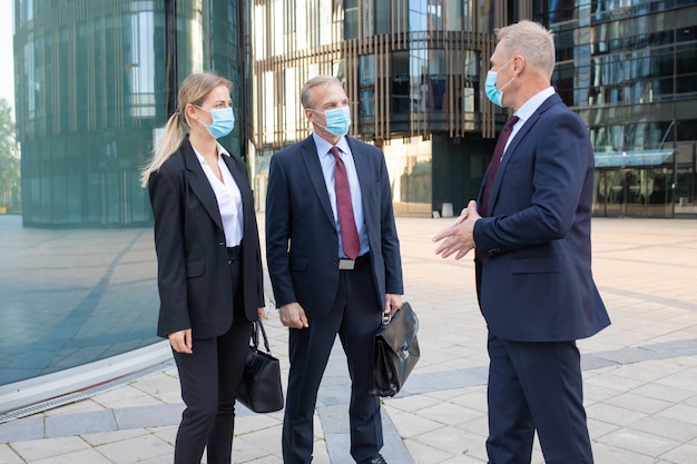 Three content business partners in masks discussing deal outside. Confident successful managers standing on street and working during coronavirus pandemic. Negotiation and partnership concept