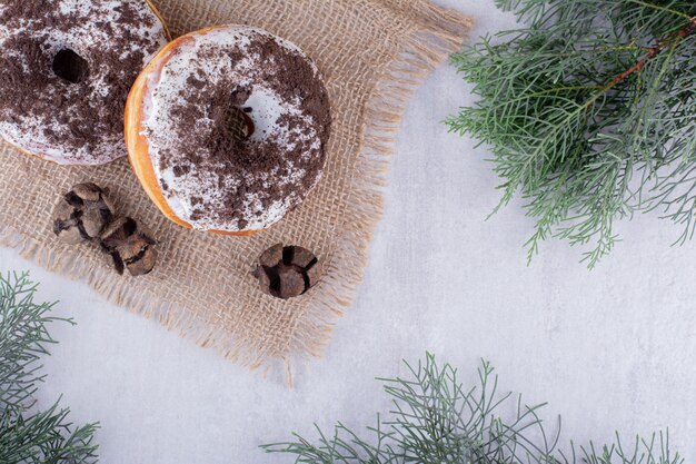 Three cypress cones and two donuts on a piece of cloth among pine branches on white background.