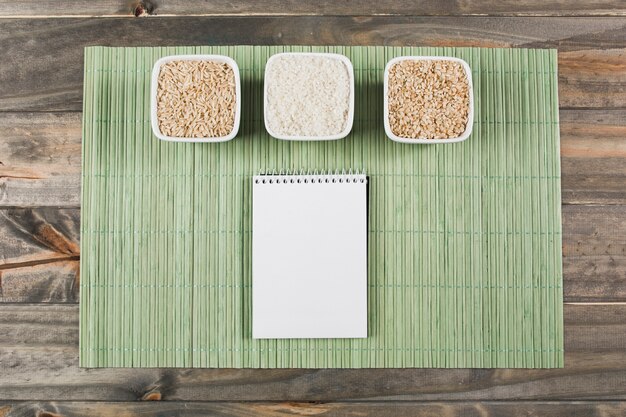 Three different type of uncooked rice bowls with spiral notepad on green placemat over the table