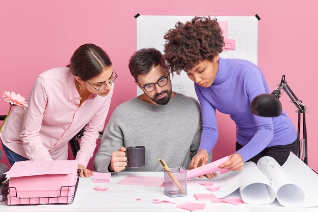 Three diverse colleague collaborate and have brainstorming meeting look attentively at papers pose at dekstop with sketches around discuss ideas of productive strategy meet in company office