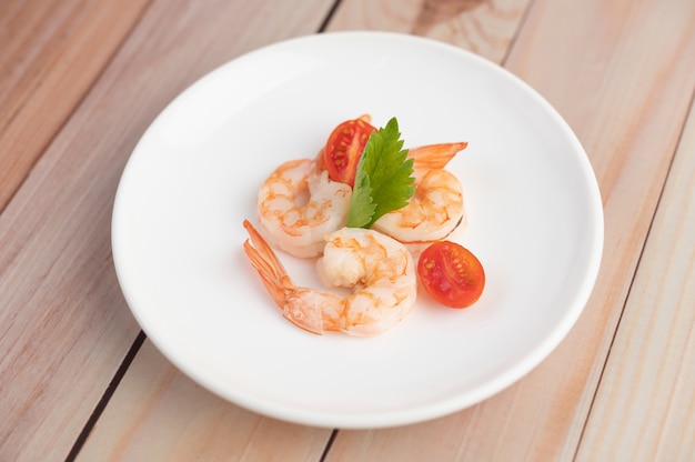 Three fresh shrimp and half tomatoes in a white plate on a wooden.