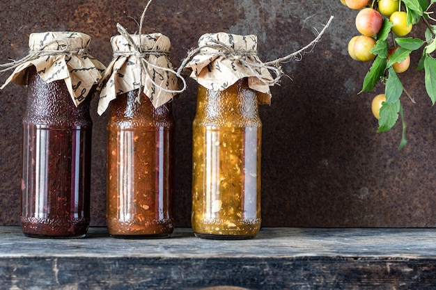 Three glass bottles of assorted Georgian tkemali sauce with ingredients on rustic wooden table.