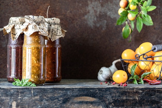 Three glass bottles of assorted Georgian tkemali sauce with ingredients on rustic wooden table.