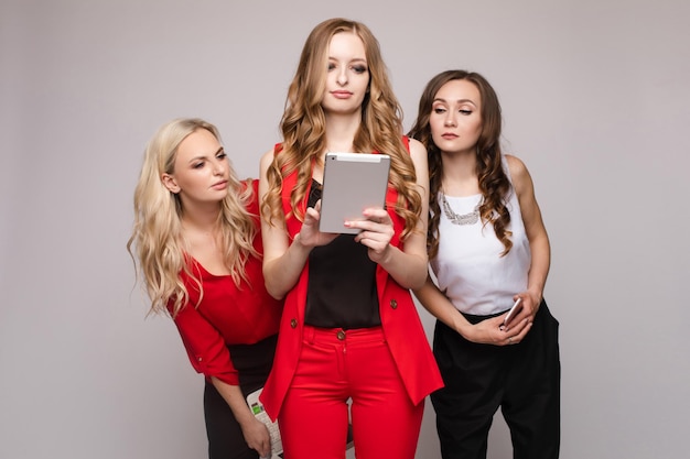 Tre splendide ed eleganti giovani donne in abiti casual che utilizzano il telefono cellulare e il tablet del computer portatile stanno usando gadget per lavoro e comunicazione