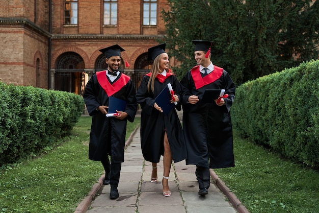 Free photo three gradutes friends in graduation gowns walking in campus.